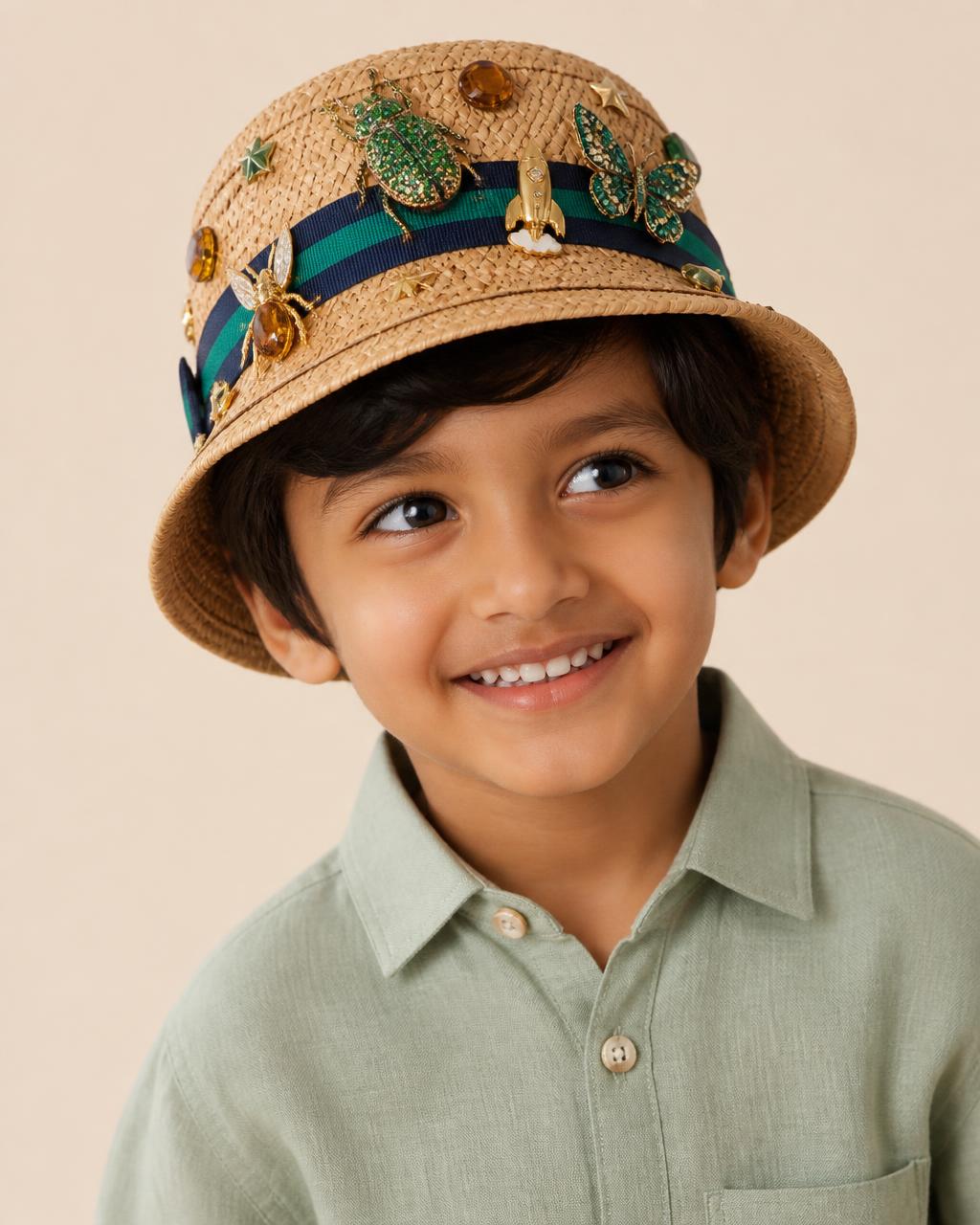 Five year old South Asian boy in a sage linen shirt wearing a small straw bucket hat with a jewelled emerald beetle, crystal butterfly, gold rocket charm and amber gems